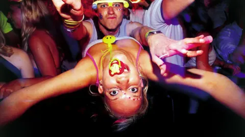 Getty Images A man wearing florescent glasses and a woman with a dummy in her mouth face the camera at a club from 2001. The picture is taken from slightly above and the woman is leaning backwards so her face is upside down