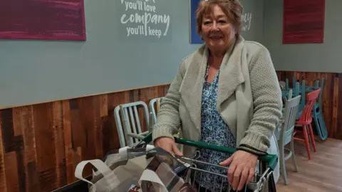 BBC A woman with brown hair and a cardigan smiles as she stands in a cafe in front of a supermarket trolley with shopping inside.