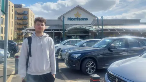 A man with black curly hair in a grey hoodie and black trousers in front of a Morrisons