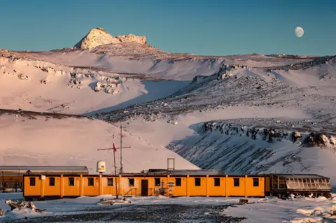 Dariusz Puczko A single storey long yellow building at the foot of snow-covered mountains. There is a blue sky behind and the moon is visible.