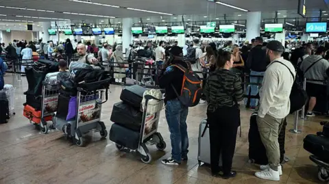 Getty Images Queues of people with trolleys filled with baggage wait at a busy airport terminal 