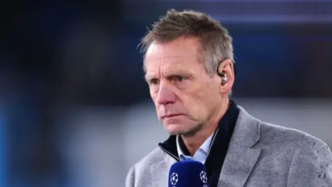 Getty Stuart Pearce, working as a pundit for Amazon Prime during the UEFA Champions League. He has short grey hair and is wearing a white shirt, dark-blue jacket and grey suit jacket. He has a blue Champion's League microphone in his hand.