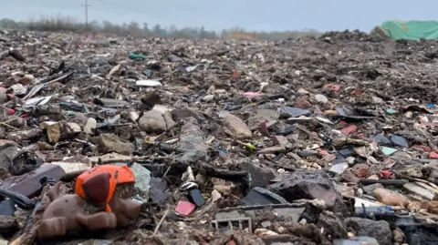 Malcolm Prior/BBC A small plastic toy dog sits on the edge of a vast waste dump, a mound of mixed rubbish which disappears into the distance where the horizon is lined with trees