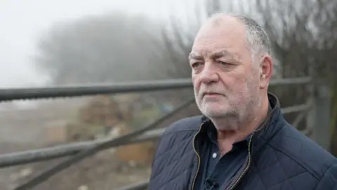 Charlie Coats stands in front of a metal gate that bars entry to the dump site. It is a grey misty winter day, with the site enveloped in cloud. In the background, in soft focus, can be seen a mound of waste.